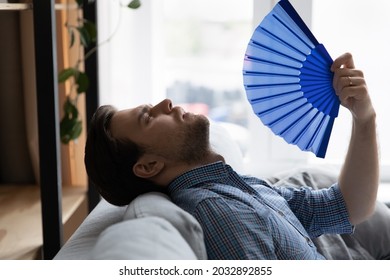 Exhausted Overheated Millennial Man Suffering From Heat At Home Without Conditioner, Trying To Cool Too Hot Air With Handheld Fan, Feeling Unwell Due To Weather, Humidity, Air Temperature, Hypoxia