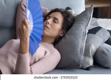 Exhausted overheated woman waving blue paper fan close up, lying on couch, relaxing on soft pillows, enjoying fresh air, tired young female suffering from heating, hot summer weather or fever