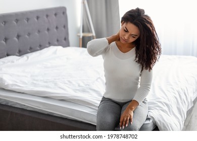 Exhausted Person. Tired African American lady sitting on bed massaging sore neck, feeling pain after sleeping on bed uncomfortable mattress, free copy space. Black woman suffering from inflammation