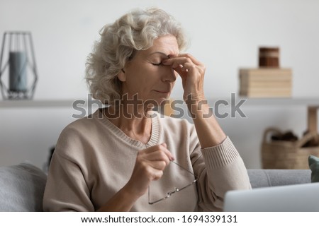 Exhausted senior older woman taking off glasses, suffering from dry eyes syndrome after computer overwork, sitting alone on sofa in living room. Middle aged grandmother rubbing eye, feeling tired.