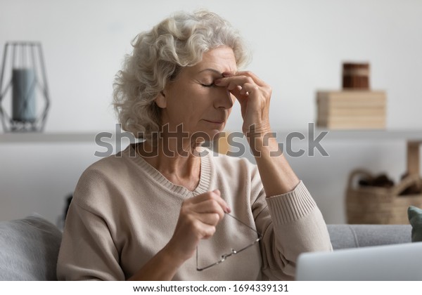 Exhausted senior older woman taking off glasses,\
suffering from dry eyes syndrome after computer overwork, sitting\
alone on sofa in living room. Middle aged grandmother rubbing eye,\
feeling tired.