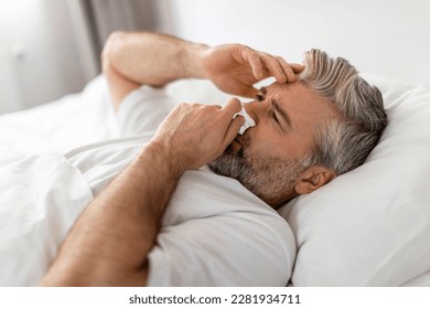 Exhausted sick middle aged grey-haired bearded man wearing pajamas lying in bed with fever and runny nose, touching head and sneezing nose, suffering from cold, flu, coronavirus, closeup