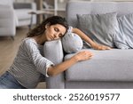 Exhausted sleepy young woman resting on floor at home, leaning head and hands on grey soft couch, enjoying leisure, break with closed eyes, accidently falling asleep near sofa