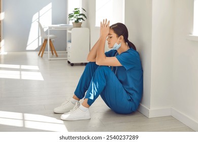 Exhausted stressed female nurse or doctor wearing blue face mask and uniform sitting with closed eyes on hospital floor in hallway. Young woman physician tired from hard work in medical clinic. - Powered by Shutterstock