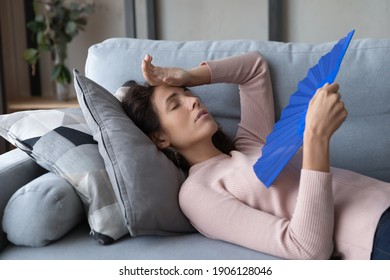 Exhausted Stressed Millennial Woman Lying On Sofa, Suffering From Hot Temperature Without Air Conditioning System, Using Paper Fan Breathing Fresh Cooled Air, Feeling Overheated Unwell Alone At Home.