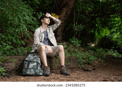 An exhausted and sweaty Asian male hiker sits on a stone, resting and taking a break during his hiking trip in the forest alone. - Powered by Shutterstock