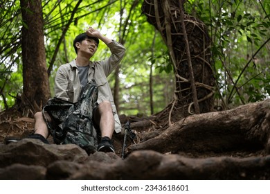 An exhausted and sweaty Asian male hiker is sitting and taking a break while hiking in the green forest. - Powered by Shutterstock