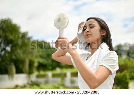 An exhausted and sweaty Asian woman in sportswear using a portable handy fan, feeling hot and tired after a long run at the park on a sunny day.