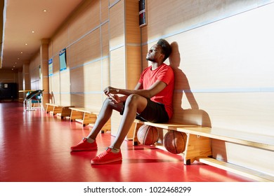 Exhausted thoughtful handsome young African-American basketball player sitting on bench with two balls under it and texting message on smartphone while stuck in training - Powered by Shutterstock