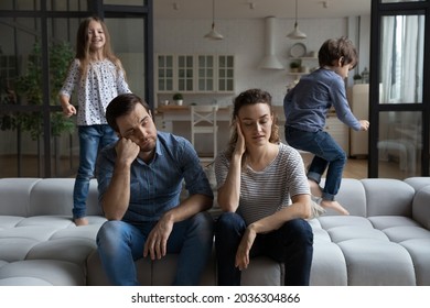 Exhausted tired couple of parents touching heads while two hyperactive noisy naughty kids jumping on couch, shouting, laughing, playing active games, having fun. Parenthood, upbringing, quarantine