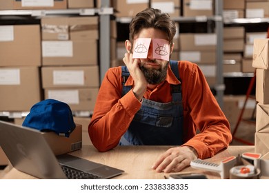 Exhausted tired warehouse worker with painted eyes on stickers, adhesive notes on face sleeping at workplace, sitting at desk with laptop, lazy young male working on difficult project. Copy space - Powered by Shutterstock