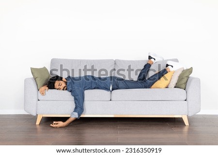 Exhausted tired young eastern man wearing casual outfit sleeping on couch at home over white blank wall. Drunk indian guy lying on sofa, experiencing hangover, copy space