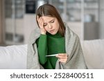 Exhausted upset woman sits on couch, feeling unwell and checking temperature with thermometer. Symptoms of illness include headache, dizziness and discomfort, typical of cold, flu season, pandemic