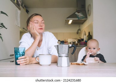 Exhausted Woman With Baby Is Sitting With Coffee In Kitchen. Modern Young Tired Mom And Little Child After Sleepless Night. Life Of Working Mother With Baby. Postpartum Depression On Maternity Leave.