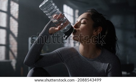 Exhausted woman in sportswear sitting box at fitness training in dark gym. Fit girl drinking water sports bottle at cardio workout. Tired athlete resting at sport club. Female bodybuilder making break