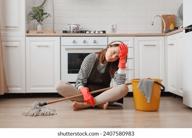 Exhausted woman wearing apron and rubber gloves housewife cleaning floor with steam mop in kitchen at home working long hours having headache sitting on floor with sad face - Powered by Shutterstock