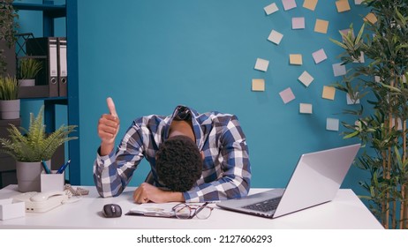 Exhausted worker putting head on desk and showing thumbs up sign, working overtime and feeling tired. Overworked man doing okay gesture with hand, waiting to take break from financial work. - Shutterstock ID 2127606293