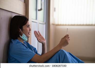 Exhausted Young Asian Woman Nurse Or Medical Worker Sitting On The Floor And Using Phone Video Call To Her Family While Taking A Break In Hospital. Overworked During Coronavirus Pandemic.