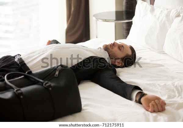 Exhausted young businessman laying with\
outstretched arms on bed next to small suitcase in hotel room.\
Entrepreneur tired after traveling, jet lag. Relieved business\
worker resting after long\
flight.