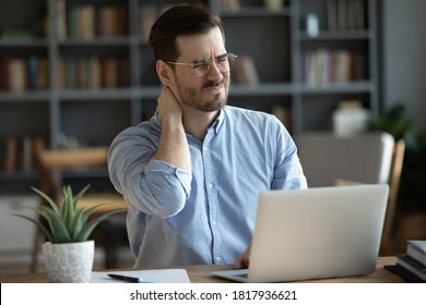 Exhausted Young Caucasian Male Worker Sit At Desk Massage Neck Suffer From Strain Spasm Muscles. Tired Unwell Man Overwhelmed With Computer Work Sedentary Lifestyle Struggle With Back Pain Or Ache.