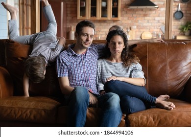 Exhausted Young Caucasian Parents Sit On Sofa At Home Annoyed By Ill-behaved Small Kids Play Have Fun. Unhappy Upset Mom And Dad Distressed Tired By Naughty Active Children. Upbringing Concept.