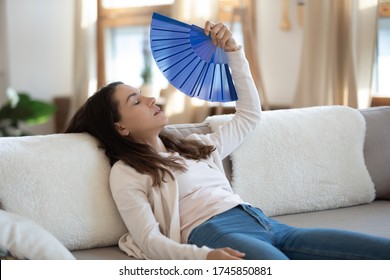 Exhausted Young Caucasian Woman Sit Relax On Couch In Living Room Wave With Hand Fan Suffer From Hot Weather, Overheated Female Use Waver Feel Unwell Unhealthy Lack Air Condition System Indoors