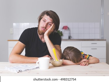 Exhausted Young Dad Sitting With His Kid On Kitchen On Early Morning And Trying To Feed 