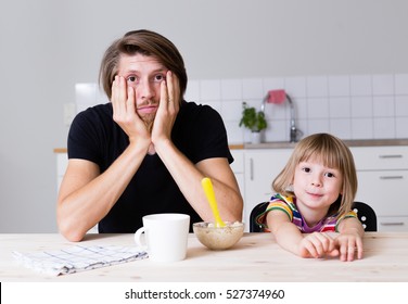 exhausted young dad sitting with his kid on kitchen on early morning and trying to feed 