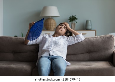 Exhausted young Indian girl feeling hot, tired, sick, resting on couch at home, waving paper handheld fan for cooking, suffering from heat attack, hypoxia, headache, touching head - Shutterstock ID 2184679443