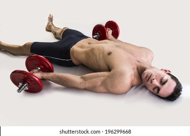 Exhausted young male bodybuilder resting on floor with dumbbells in hands