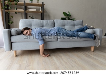 Exhausted young man fell asleep on comfortable couch in modern living room, having no energy after hard working day. Tired depressed unmotivated caucasian guy napping on sofa at home, fatigue concept.