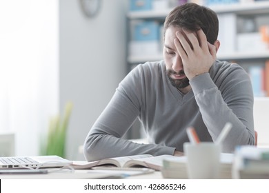 Exhausted young man working at office desk and touching his head, he is having a bad headache, stress and overwork concept