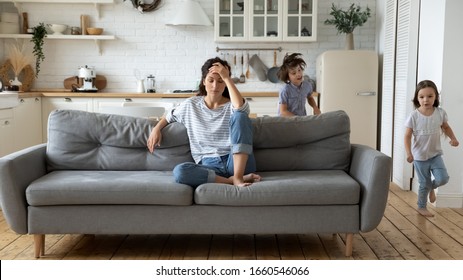 Exhausted Young Mum Sit On Couch In Kitchen Feel Unwell Tired From Ill-behaved Loud Little Children Running Playing, Sick Annoyed Mother Or Nanny Relax On Sofa Suffer From Headache, Parenting Concept