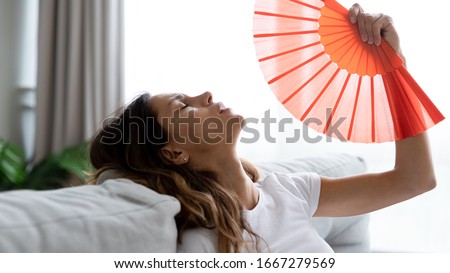 Exhausted young woman sit relax on couch in living room waving with hand fan suffer from hot temperature summer heat at home, sick millennial girl use waver struggle with heatstroke lack fresh air