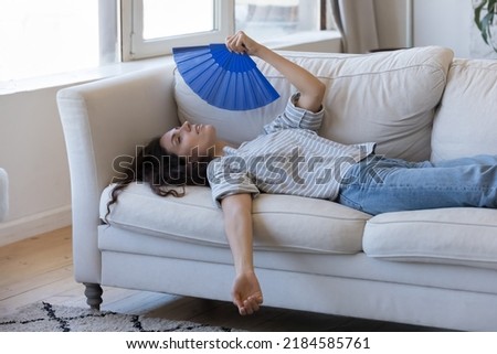 Exhausted young woman suffering from hot weather, overheat, too high air temperature at home, lying on sofa, waving paper handheld fan, cooling, feeling discomfort