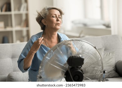 Exhauster older retired woman sitting at electric fan with propeller, enjoying cool fresh air flow, feeling relief, sitting on home sofa, suffering from hot weather, overheating