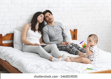 Exhausting Parenthood. Tired Millennial Parents Sitting On Bed And Want To Sleep, Active Baby Playing All Night Long