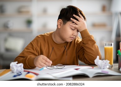 Exhautsed Chubby Asian Kid Teen Boy Doing Homework Alone, Sitting At Table Full Of Exercise Books, Notepads, Glass Of Orange Juice, Touching His Head, Experiencing Difficulties With Studying