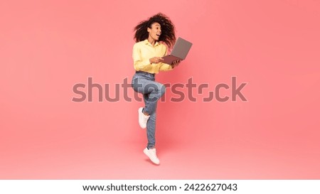 Exhilarated black woman jumping with excitement while holding laptop, her joy radiating against vibrant pink background, illustrating technology enthusiasm and success