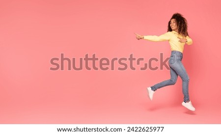 Exhilarated black woman leaps with joy, pointing aside at copy space beside her on vibrant pink background, full length shot, showcasing place and promoting something