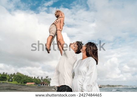 Exhilarating family moment by the sea: Father lifts joyful toddler high into the air, with mother smiling beside them, all against a dramatic sky and tranquil sea, evoking happiness and family unity