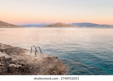 The Exit Horizon Framework - Entry point - Stone pier with metal ladder descending into calm Mediterranean water at sunset, symbolizing the decisive entry point in a long-term wealth strategy. Visual metaphor for capital deployment, strategic investment action and the transition from planning to execution in an exit strategy framework.