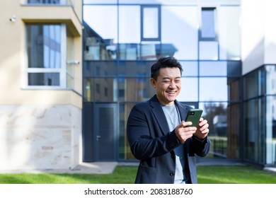 Exited Asian Businessman Celebrating Achievement Looking At Smartphone Outdoor. Successful Business Man Celebrating Win With Mobile Phone Outside. Happy Good News. Successful Deal, Startup Bet