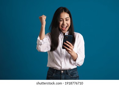 Exited Asian Girl Making Winner Gesture While Using Smartphone Isolated Over Blue Background