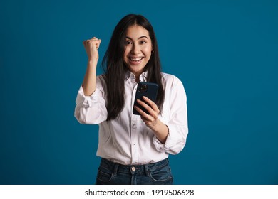 Exited Asian Girl Making Winner Gesture While Using Smartphone Isolated Over Blue Background