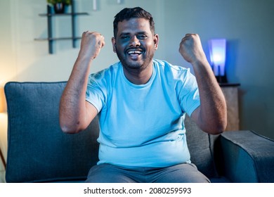 Exited Fat man watching sports on TV at night while sitting at home - Concept of cricket game supporter, watching live streaming match and weekend leisure activities.