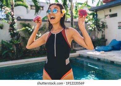 Exited slim lady in swimwear and sunglasses standing with raised arms with pitaya near swimming pool at tropical resort on summer day - Powered by Shutterstock