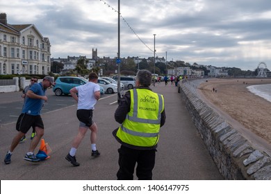 Exmouth, Devon, UK, April, 13, 2019: Running Taking Part In A Park Run Event Along The Sea Front At Exmouth, Volunteer In The Foreground Making Sure The Runners Are Ok