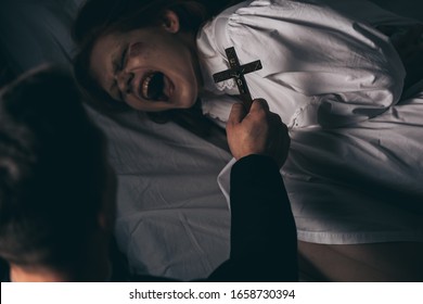 exorcist holding cross over demonic yelling girl in bed - Shutterstock ID 1658730394