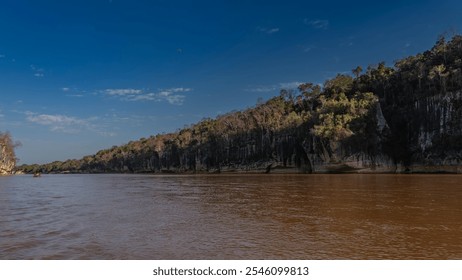 The exotic African red-brown river flows calmly. A tourist canoe can be seen in the riverbed. There is green vegetation on the steep banks. Blue sky, clouds. Madagascar. Manambolo River - Powered by Shutterstock